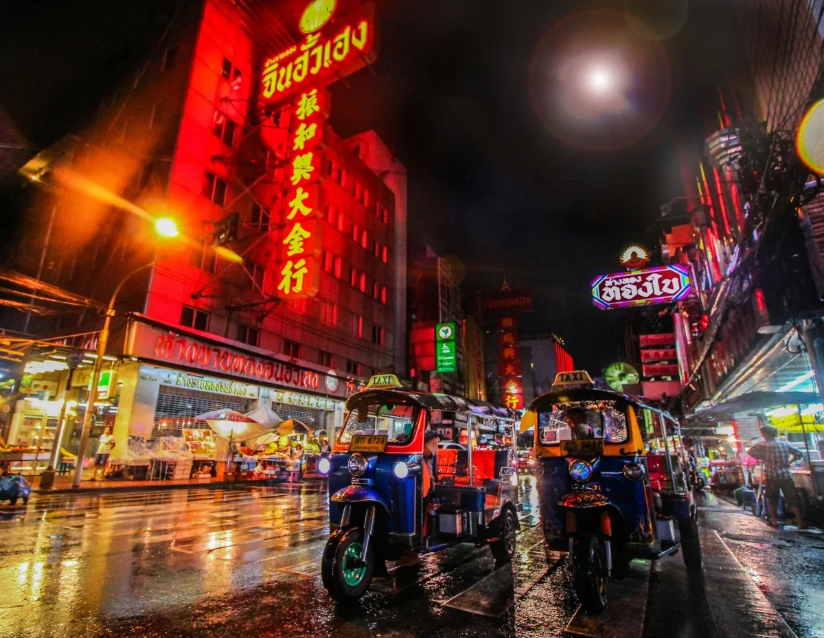 Bangkok's Chinatown (Yaowarat) at night with vibrant red and yellow neon signs and colorful tuk-tuks on wet street